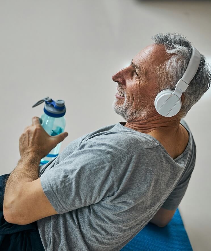 A man resting on a yoga mat wearing headphones and holding a water bottle